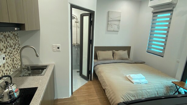 Een kleine slaapkamer met een bed, een raam met zonwering, een kitchenette met een spoelbak en een douche.