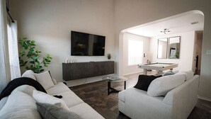 Een moderne woonkamer met een modulaire bank, een flatscreen televisie, een eettafel en een keuken met een spoelbak en spiegel.
