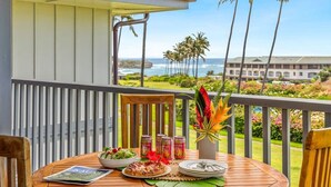 Un tavolo di legno con un pasto, una rivista e un'elegante composizione floreale tropicale su un balcone con vista su una spiaggia e l'oceano.