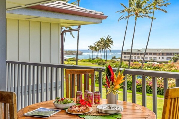 A wooden table with a meal, a magazine, and a tropical flower arrangement on a balcony overlooking a beach and ocean.