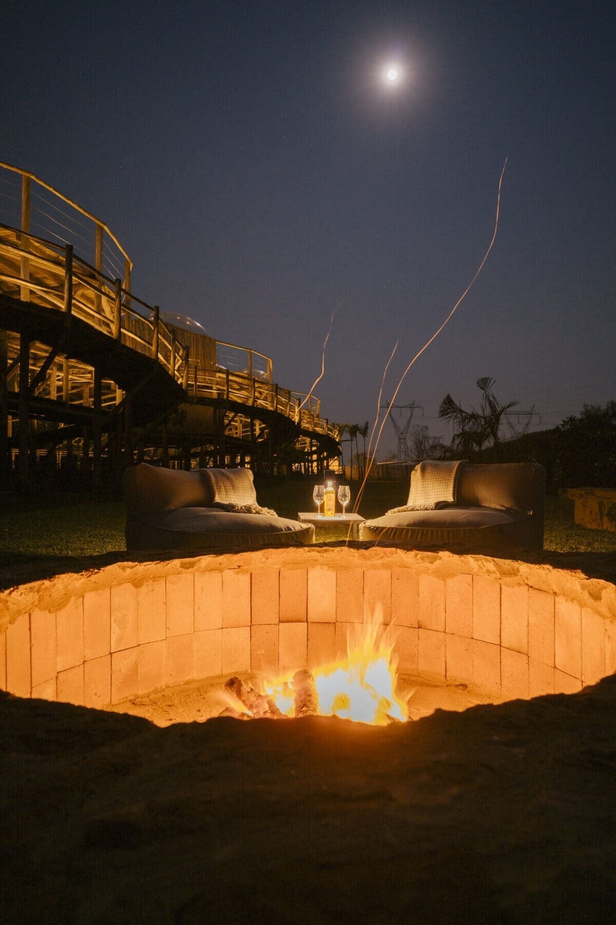A nighttime outdoor seating area with a fire pit, two sofas, and a table with glasses.