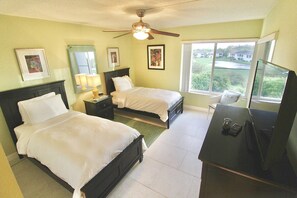 A hotel room with two beds, a desk with a TV, a ceiling fan, and a window with a view of houses.