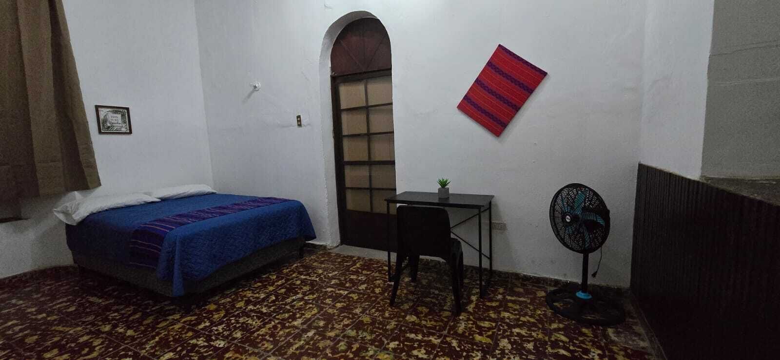 A room with a bed, a desk, a chair, a fan, and a window.