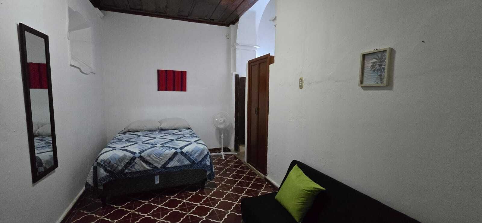A narrow room with a bed, a chair, a small table, and a fan.
