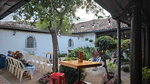 Ein Terrassenbereich im Freien mit weißen Plastikstühlen und -tischen, umgeben von Grünpflanzen und einem Gebäude mit Fenstern.