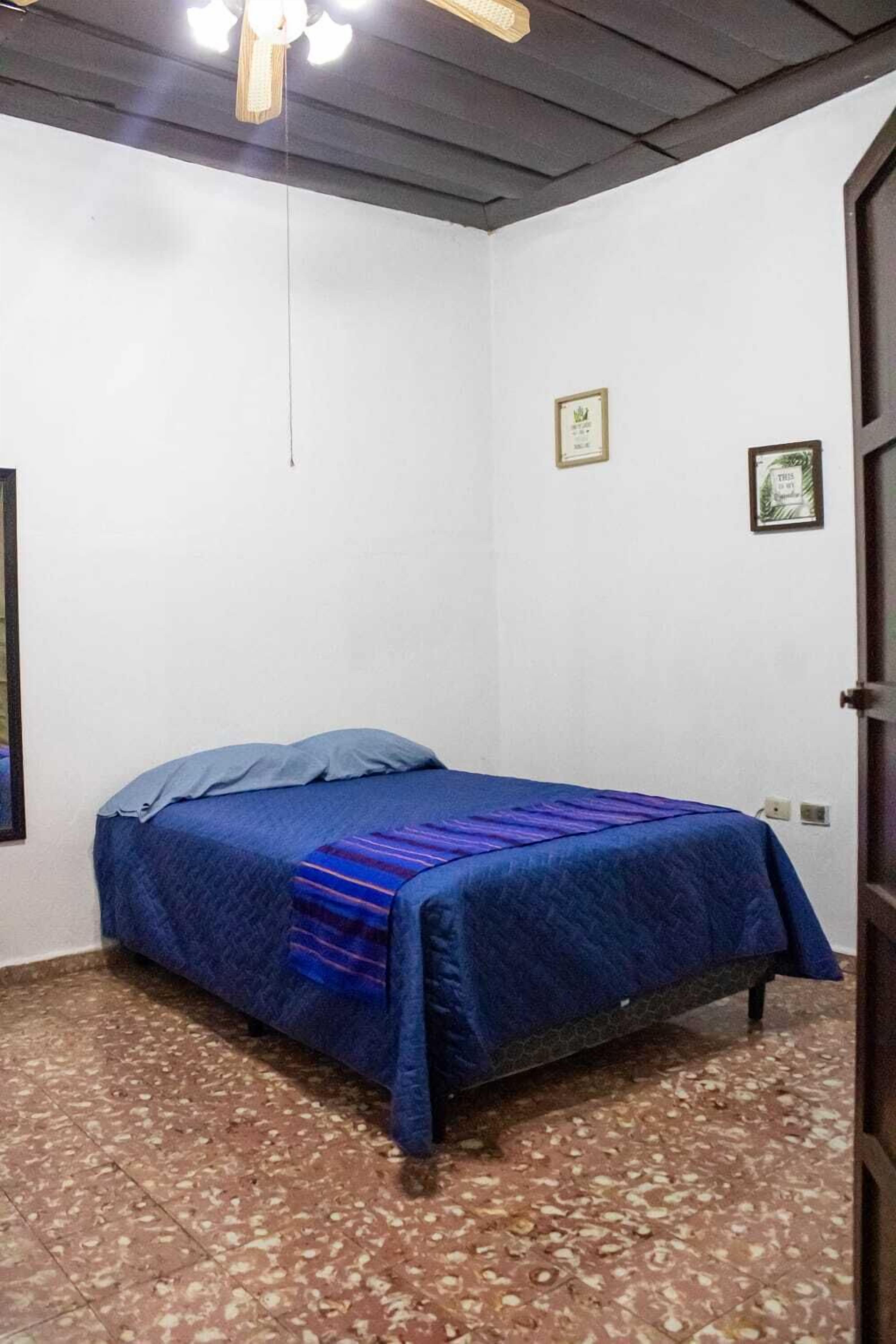A small, single-bed room with a wooden ceiling fan, white walls, and a tiled floor.