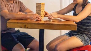 Zwei Personen spielen ein Brettspiel an einem Holztisch mit roten Hocker.