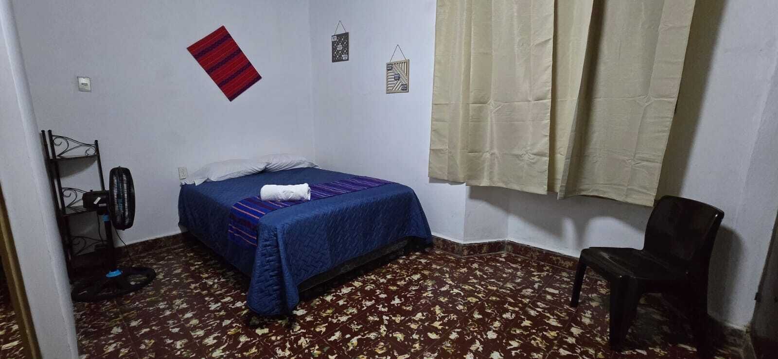 A small, single-bed room with a patterned floor, a bed with a blue blanket, a standing fan, a black chair, and a window with curtains.