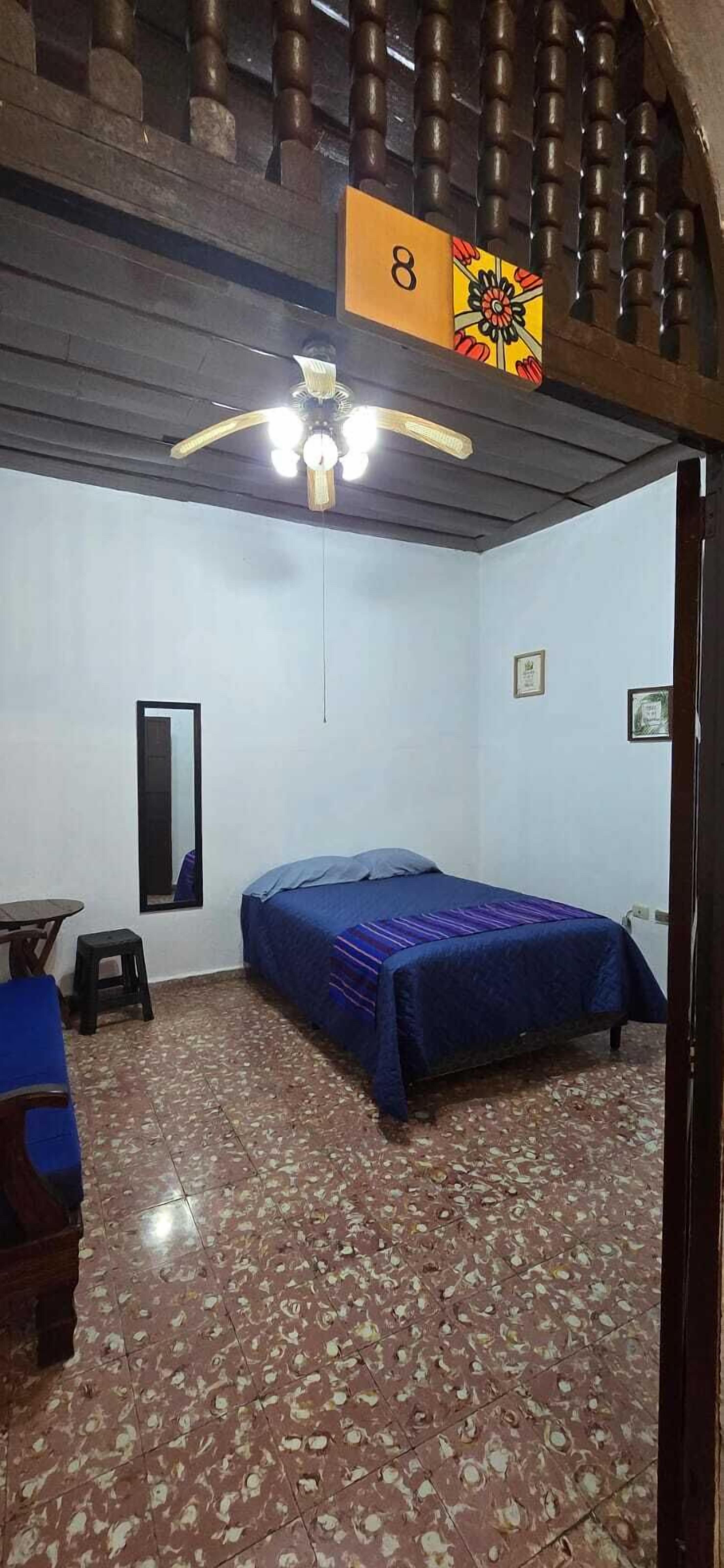 Una habitación con una cama, un ventilador de techo, una mesita y un espejo.