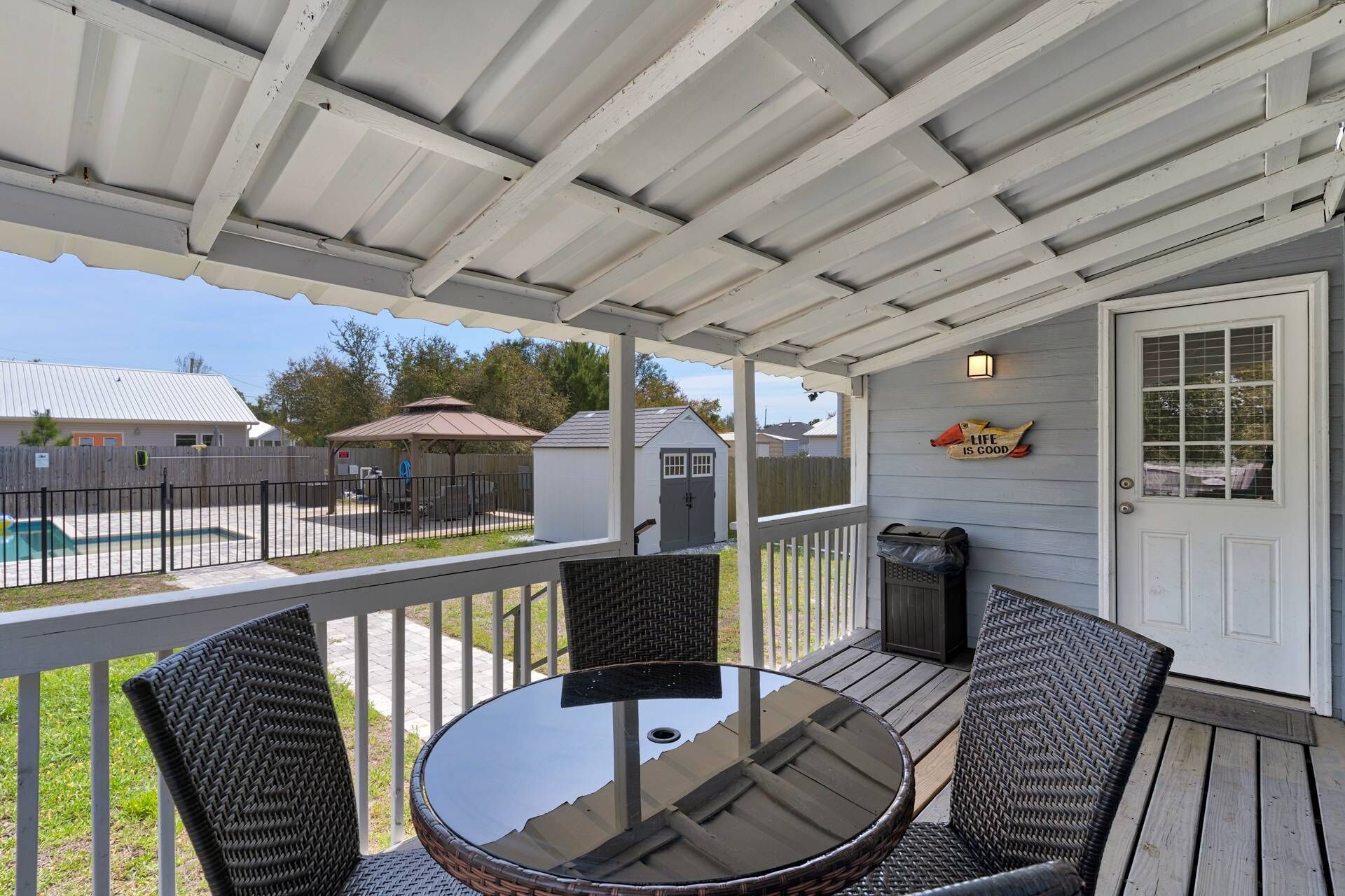 A covered patio with a round glass table and wicker chairs, a white door with a 'Life is Good' sign, and a view of a backyard with a pool and a shed.