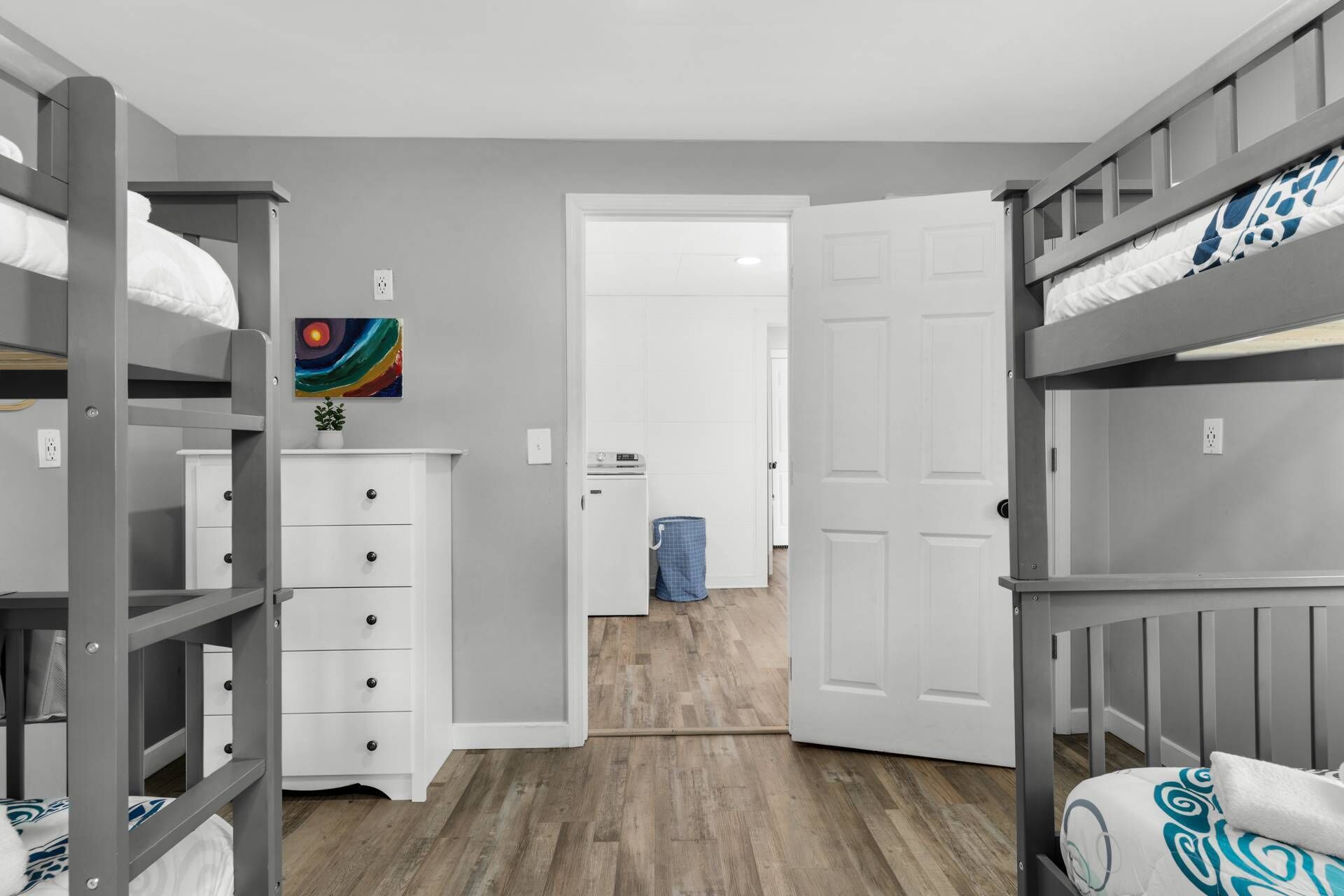 A room with bunk beds, a white dresser, a colorful painting, and a laundry area.