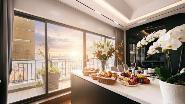 A modern kitchen with a large window overlooking a cityscape, featuring a breakfast buffet with pastries, fruits, and flowers. - Crescendo Unique Studio (Hanoi)