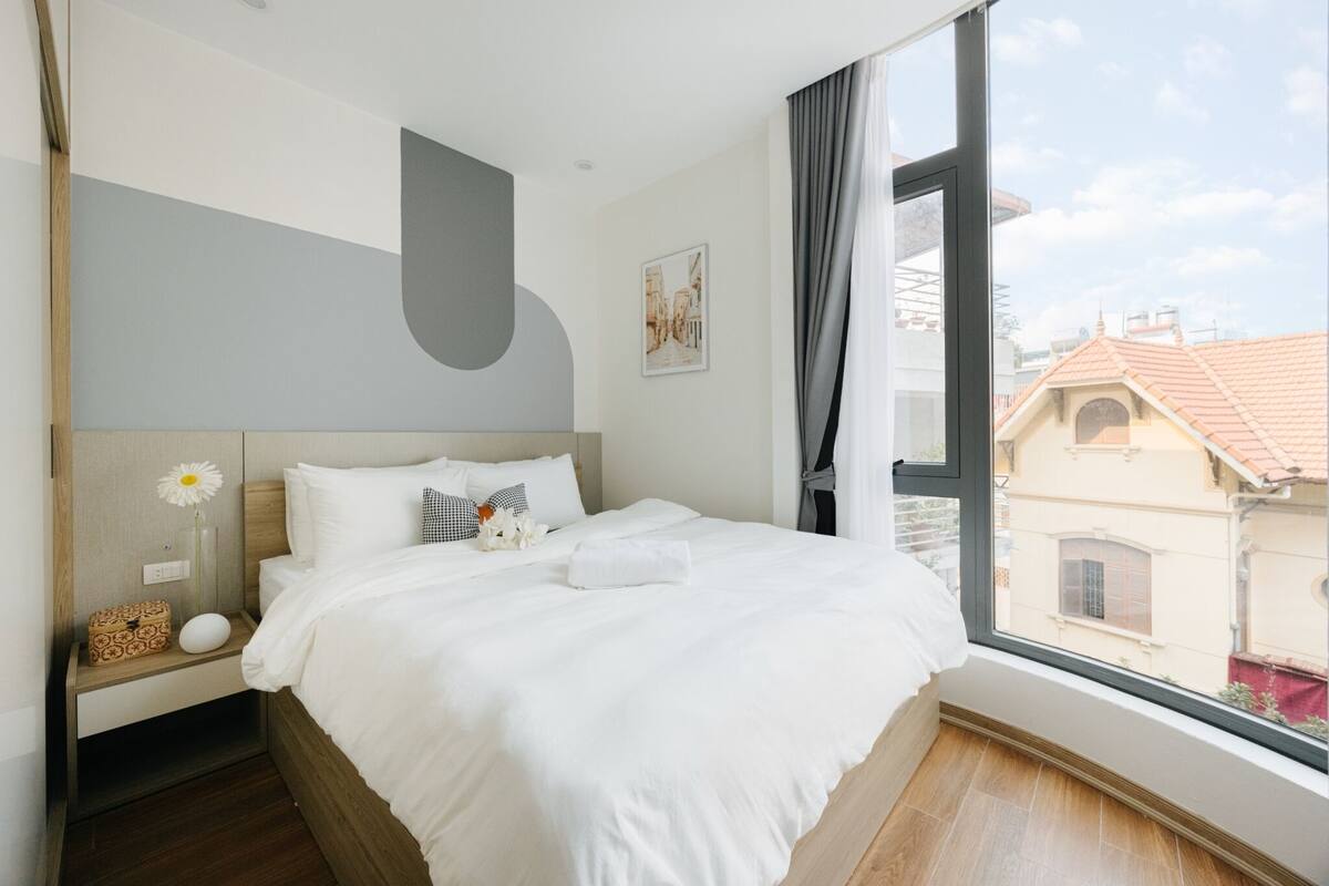 Ein modernes Hotelzimmer mit einem großen Bett, einem Nachttisch mit einer Vase und Blick auf Gebäude durch ein Fenster.