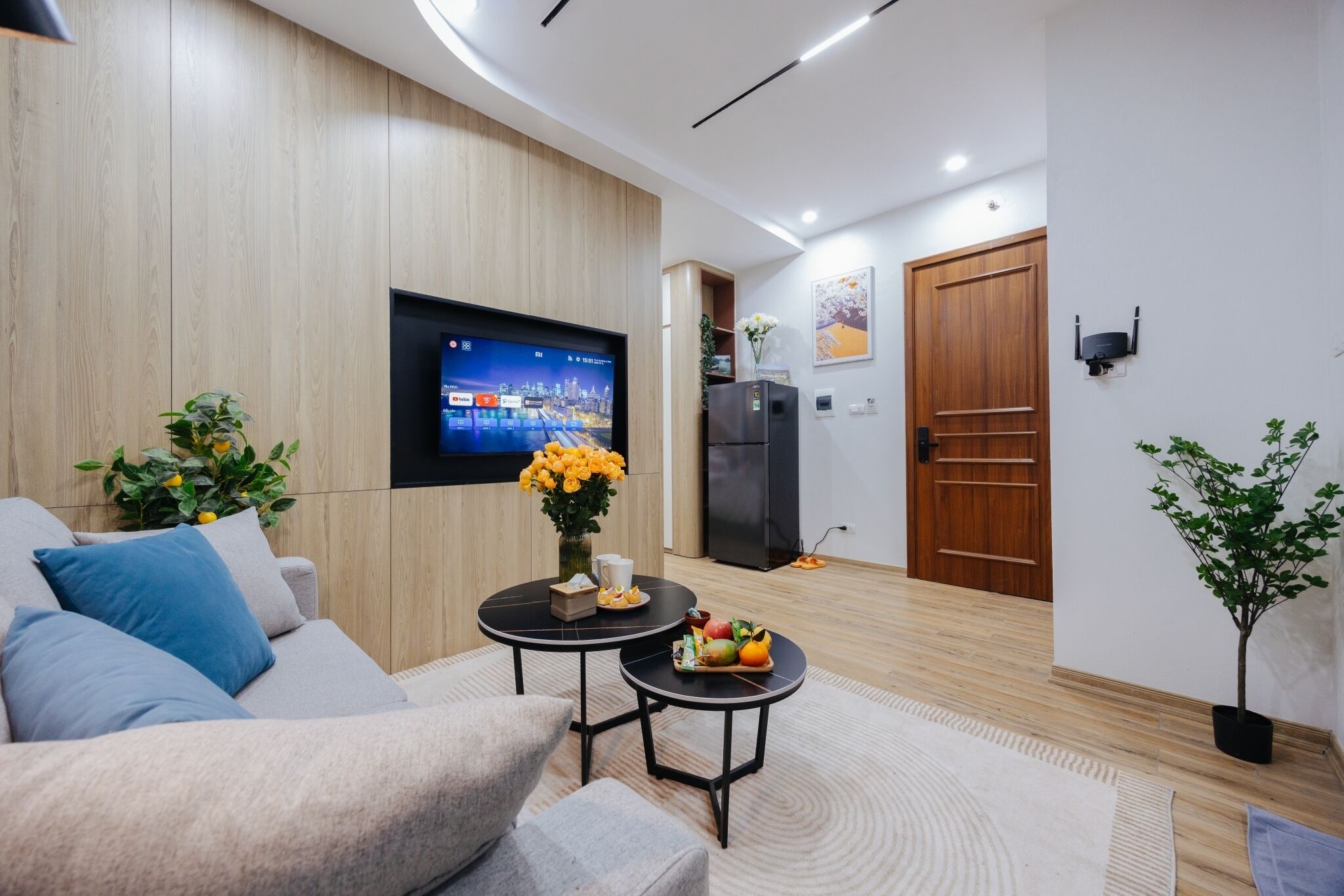 A modern living room with a sofa, two small tables, a flat-screen TV, a refrigerator, and a door.