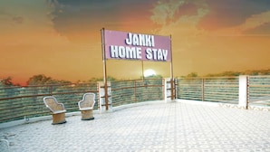 Une enseigne indiquant « JANKI HOME STAY », avec un sol à motifs et deux chaises en osier.