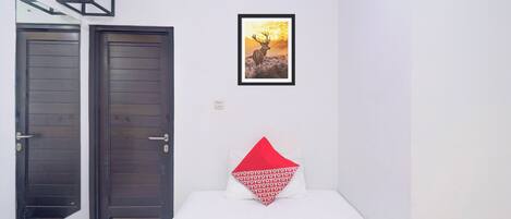A room with two doors, a bed with a red pillow, and a framed picture on the wall.
