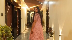 Une femme vêtue d’une robe rose marche dans un couloir dont le sol est en marbre, orné de plantes en pot et éclairé par des luminaires modernes.