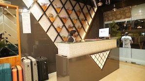A reception desk with a modern design, featuring a geometric wall pattern and a wooden panel with floral decorations. There are several suitcases placed near the desk, and a person is visible behind it. - Hotel Banaras Palace (Varanasi)