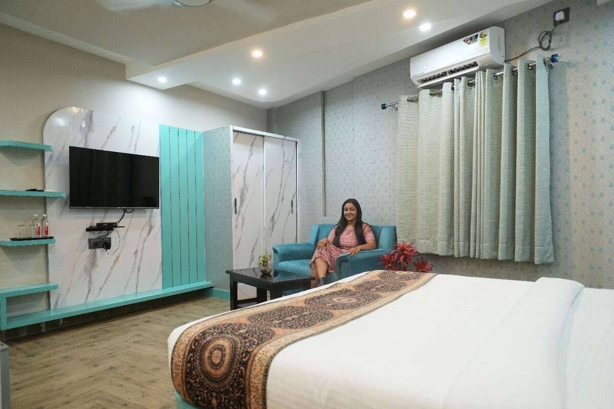 A hotel room with a bed, a seating area, a TV mounted on the wall, and an air conditioning unit.