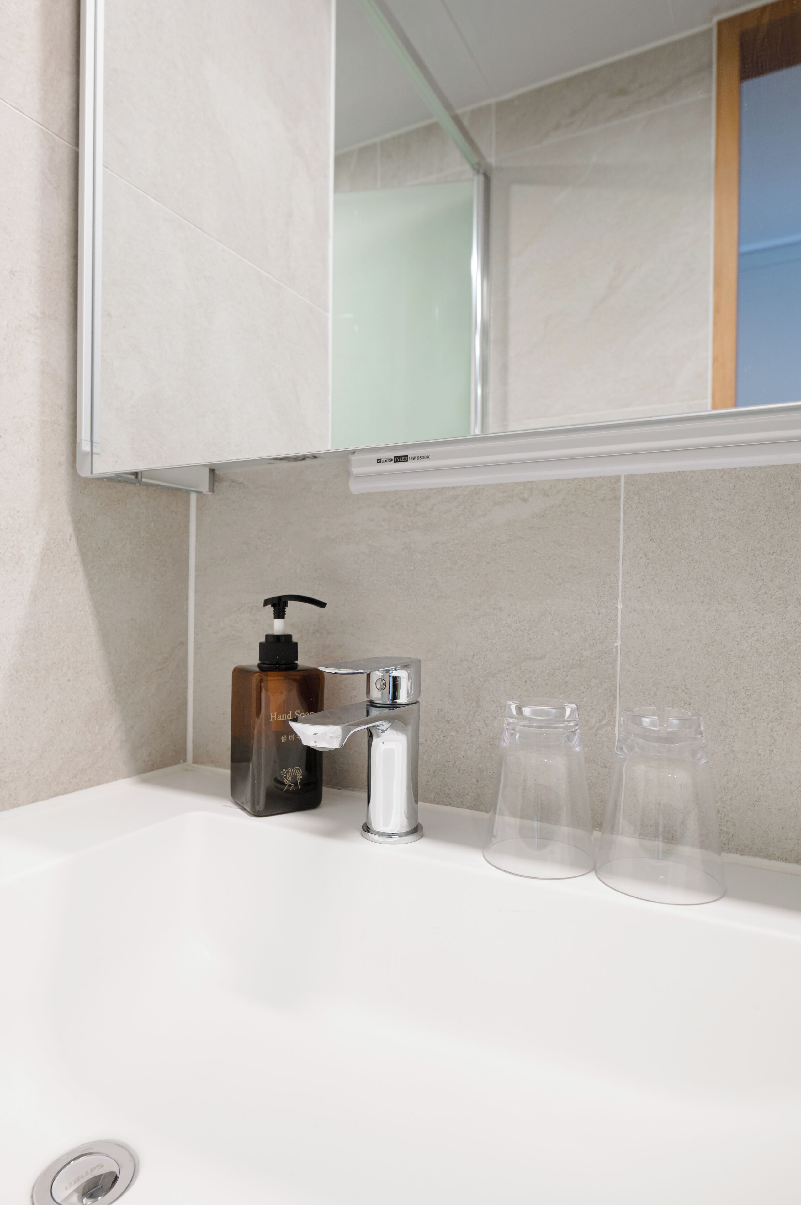 Une salle de bain avec un lavabo blanc, un miroir au-dessus, un distributeur de savon liquide et deux bouteilles en verre transparent posées sur le comptoir.