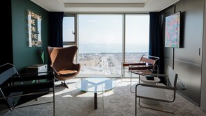 Ein modernes Hotelzimmer mit großem Fenster und Blick auf die Stadt, einem Glastisch, Ledersesseln und einem flachbildfernseher an der Wand.