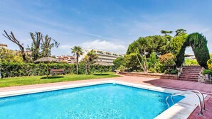 Ein Schwimmbecken mit klarem, blauem Wasser, umgeben von einem Garten mit verschiedenen Pflanzen und Bäumen, und einer Treppe, die zu einem Gebäude im Hintergrund führt.