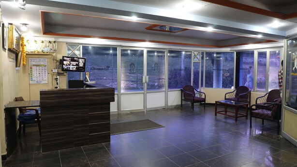 A reception area with a wooden reception desk, a seating area with chairs, a large window with a view, and a wall-mounted television. - Royal Hotels, Kodaikanal (Kodaikanal)