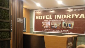 A hotel reception desk with a wooden counter and a large sign displaying the hotel name and contact information. - Hotel Indriya (Greater Noida)