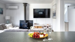 Un salon moderne avec une cheminée, un téléviseur à écran plat et une table à manger agrémentée d’un plateau de fruits et de deux verres de vin.
