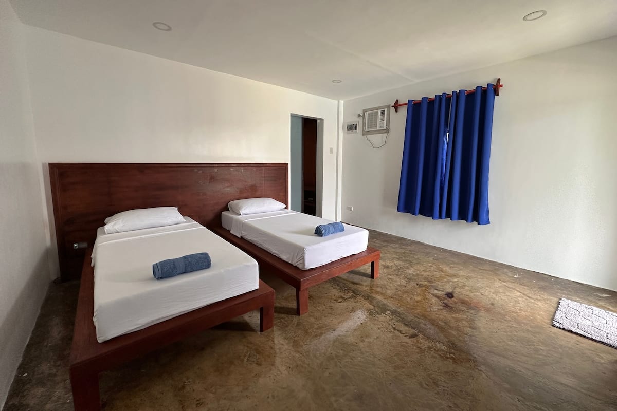 Habitación de hotel con dos camas individuales, cabecero de madera, cortina azul y una mesita.