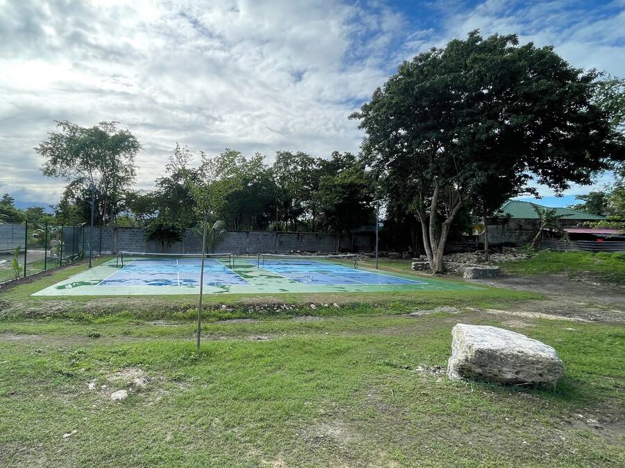 An outdoor tennis court surrounded by a green fence, trees, and a stone wall.