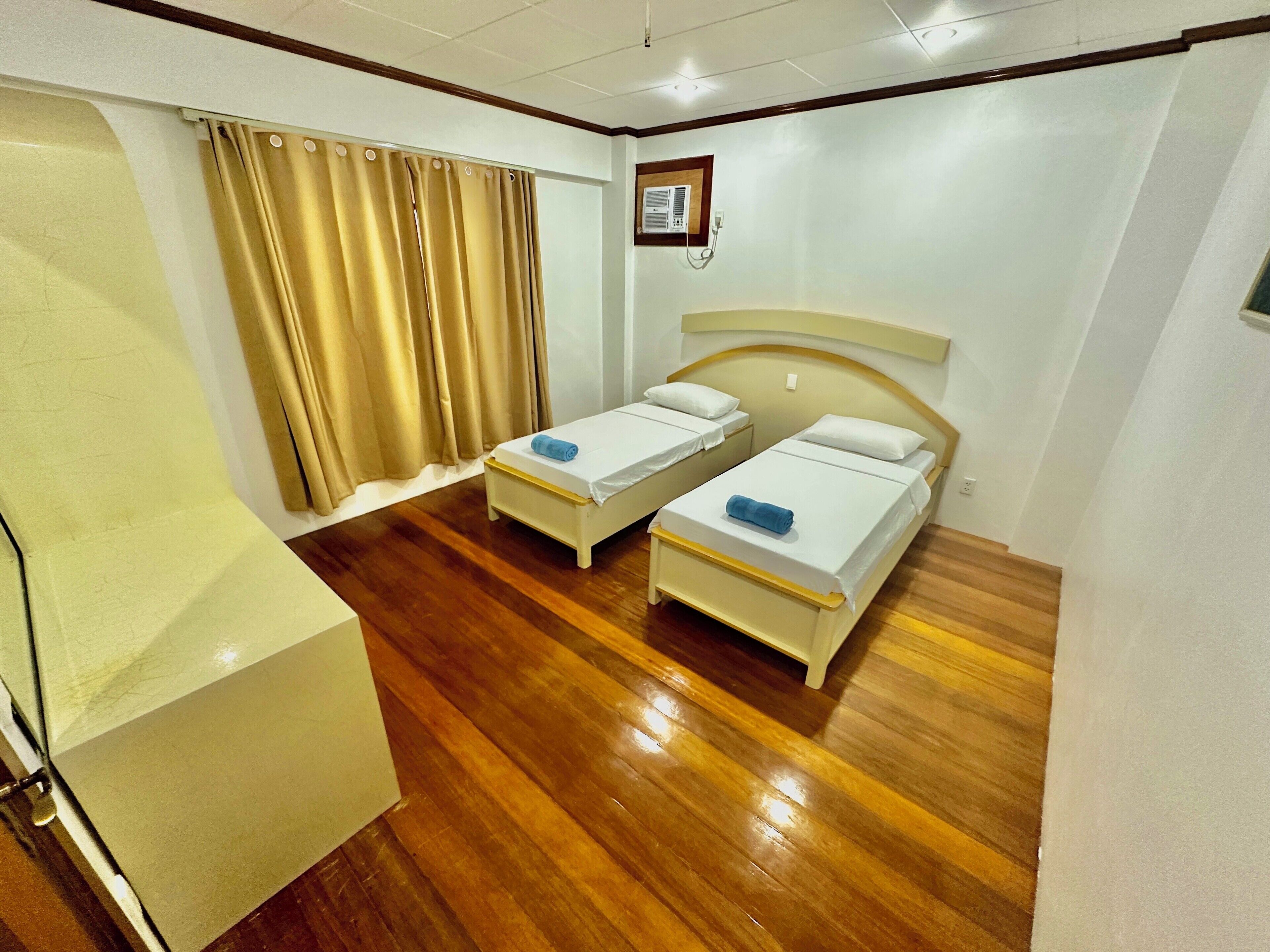 A hotel room with two beds, a wooden floor, a window with curtains, and a small air conditioning unit on the wall.