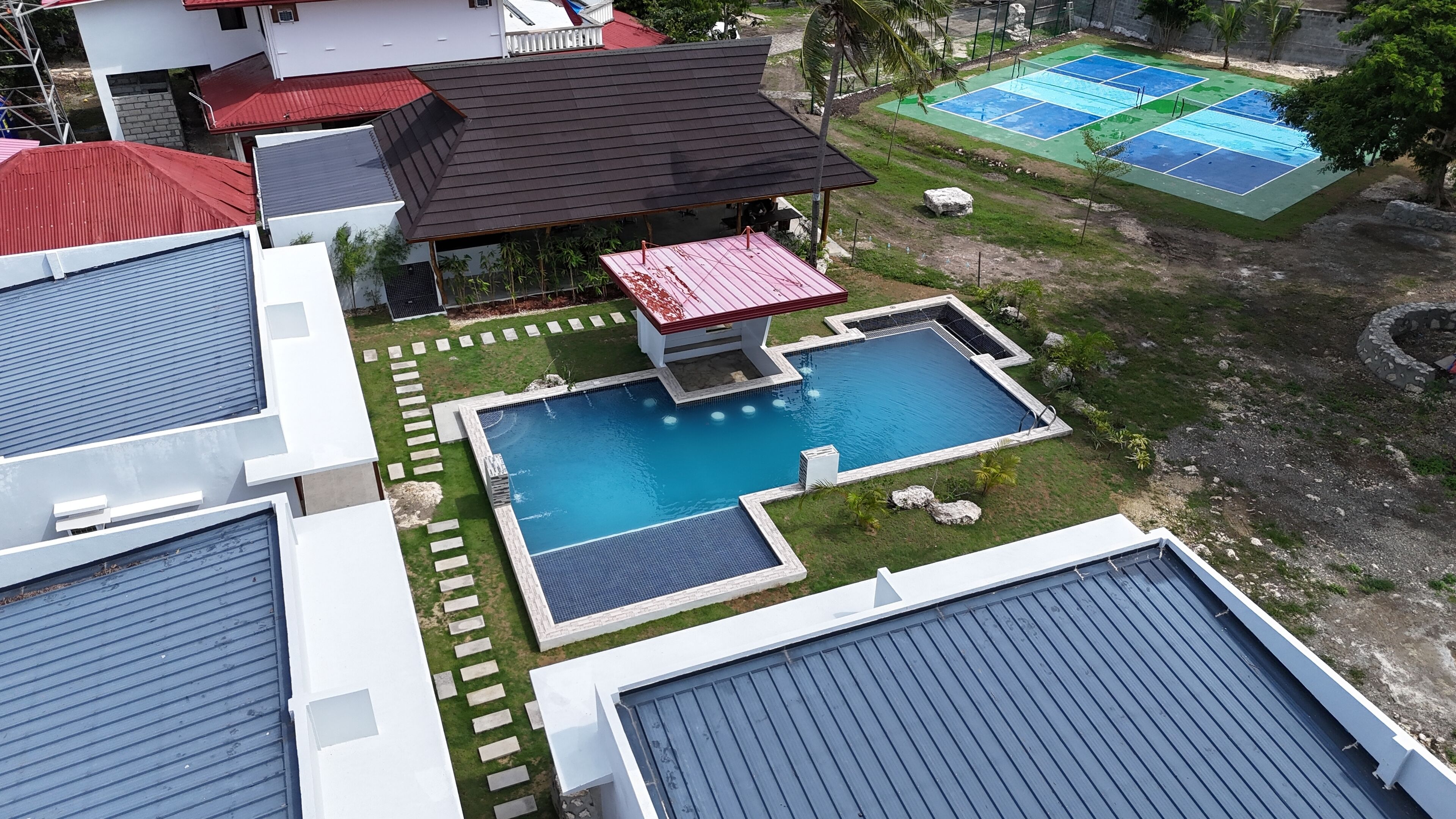 A residential area with a swimming pool surrounded by a deck, a tennis court, and a covered pavilion.