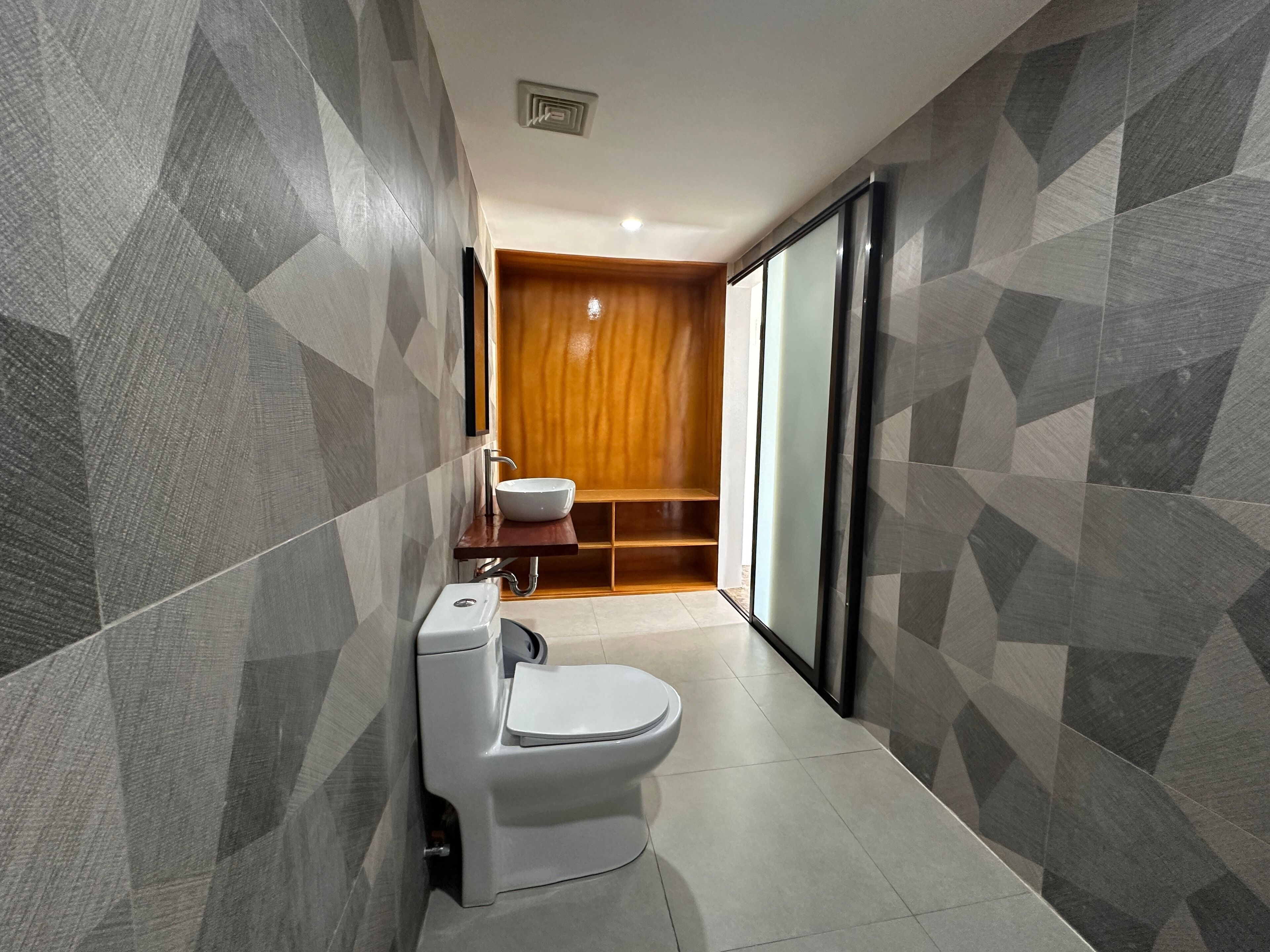A modern bathroom with a white toilet, a wooden vanity, and a geometric-patterned wall.