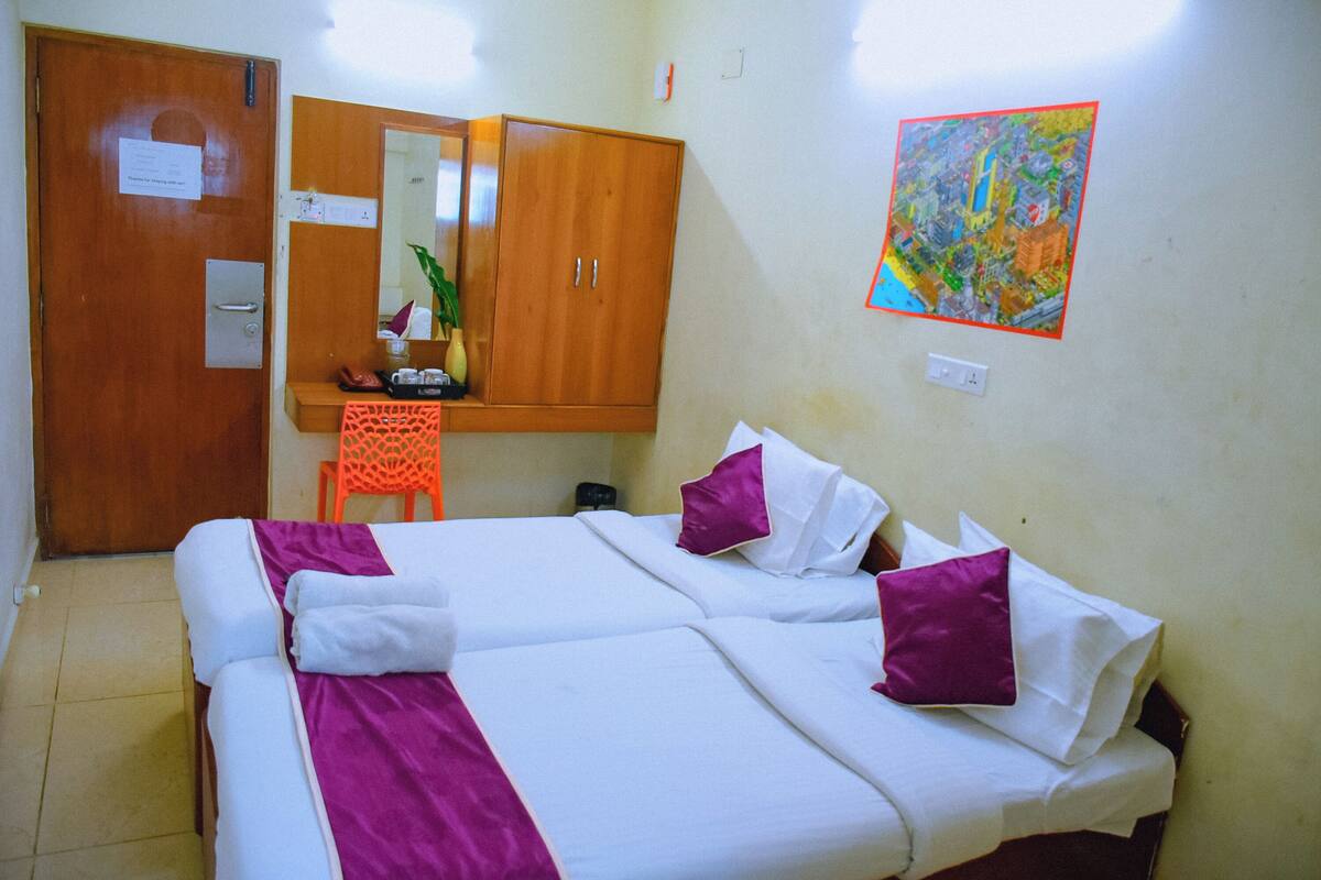 A double bed with white and purple bedding, a wooden wardrobe, a small desk with a chair, and a colorful painting on the wall.