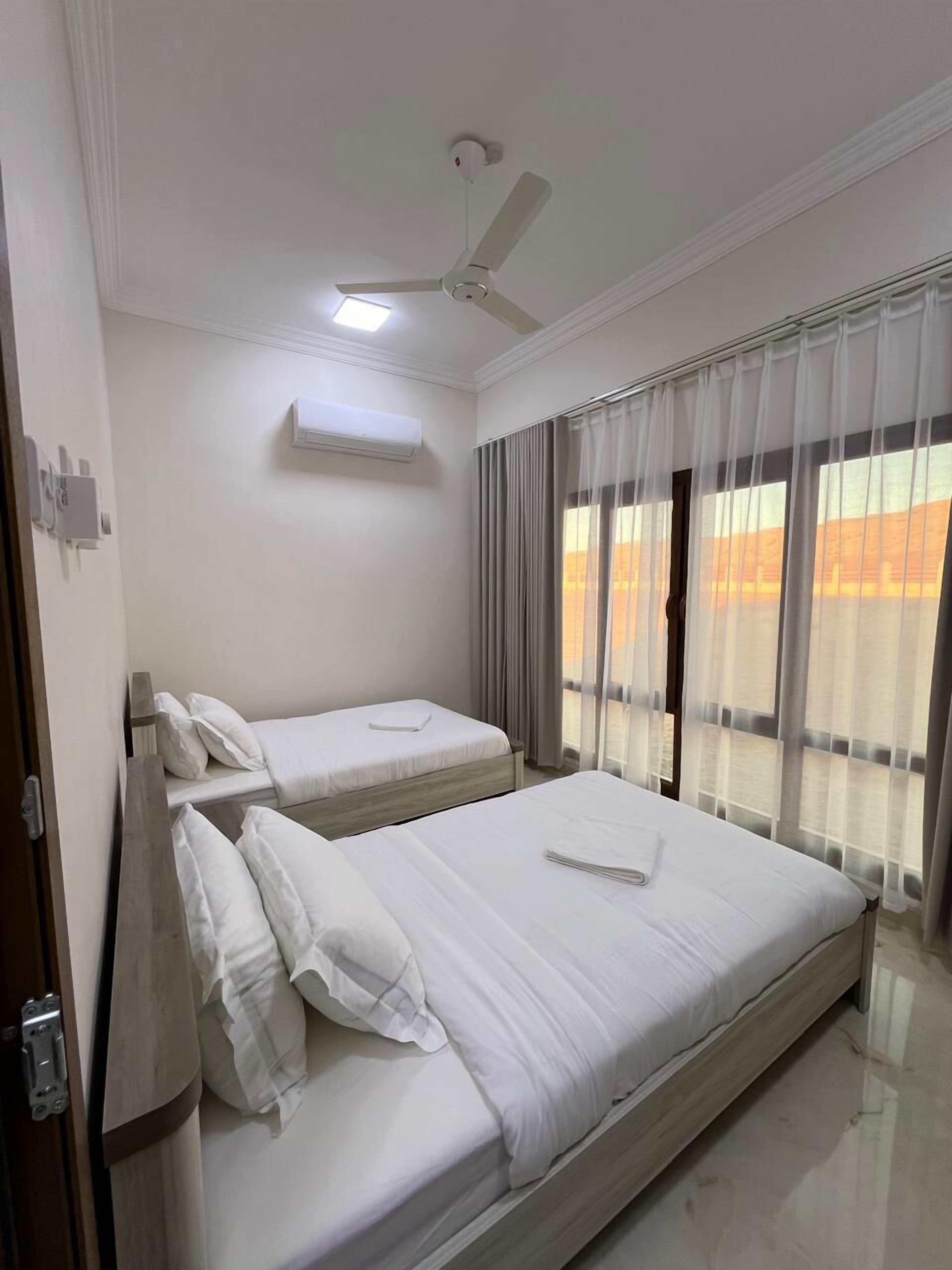 A hotel room with two beds, an air conditioning unit, a ceiling fan, and a view of the sunset through sheer curtains.