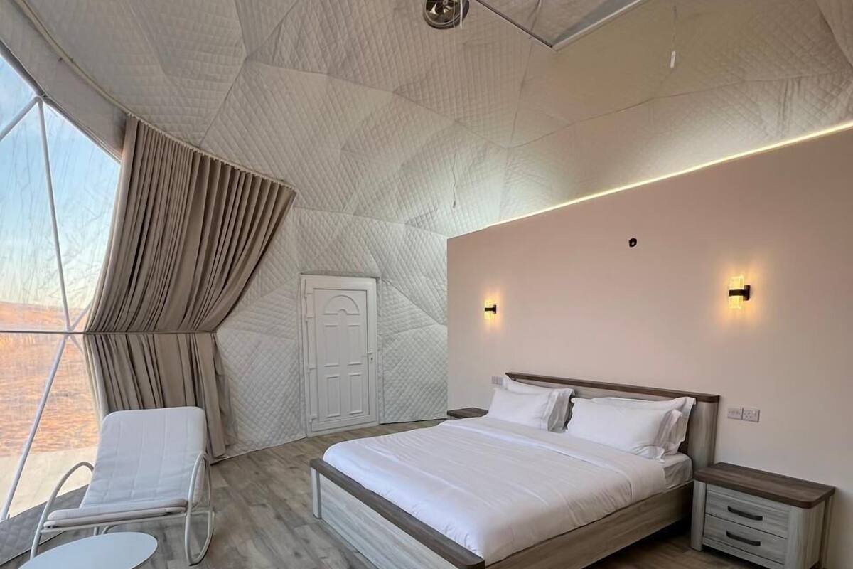 A modern bedroom with a large bed, a seating area with a table, and a view through a window.