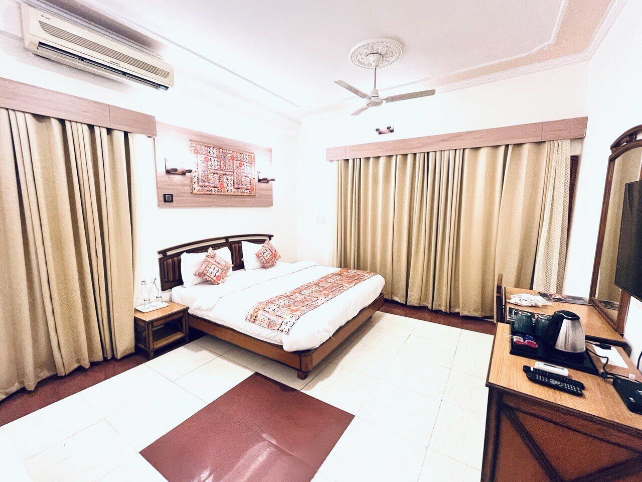 A hotel room with a bed, a desk with a computer, an air conditioner, a ceiling fan, and curtains.
