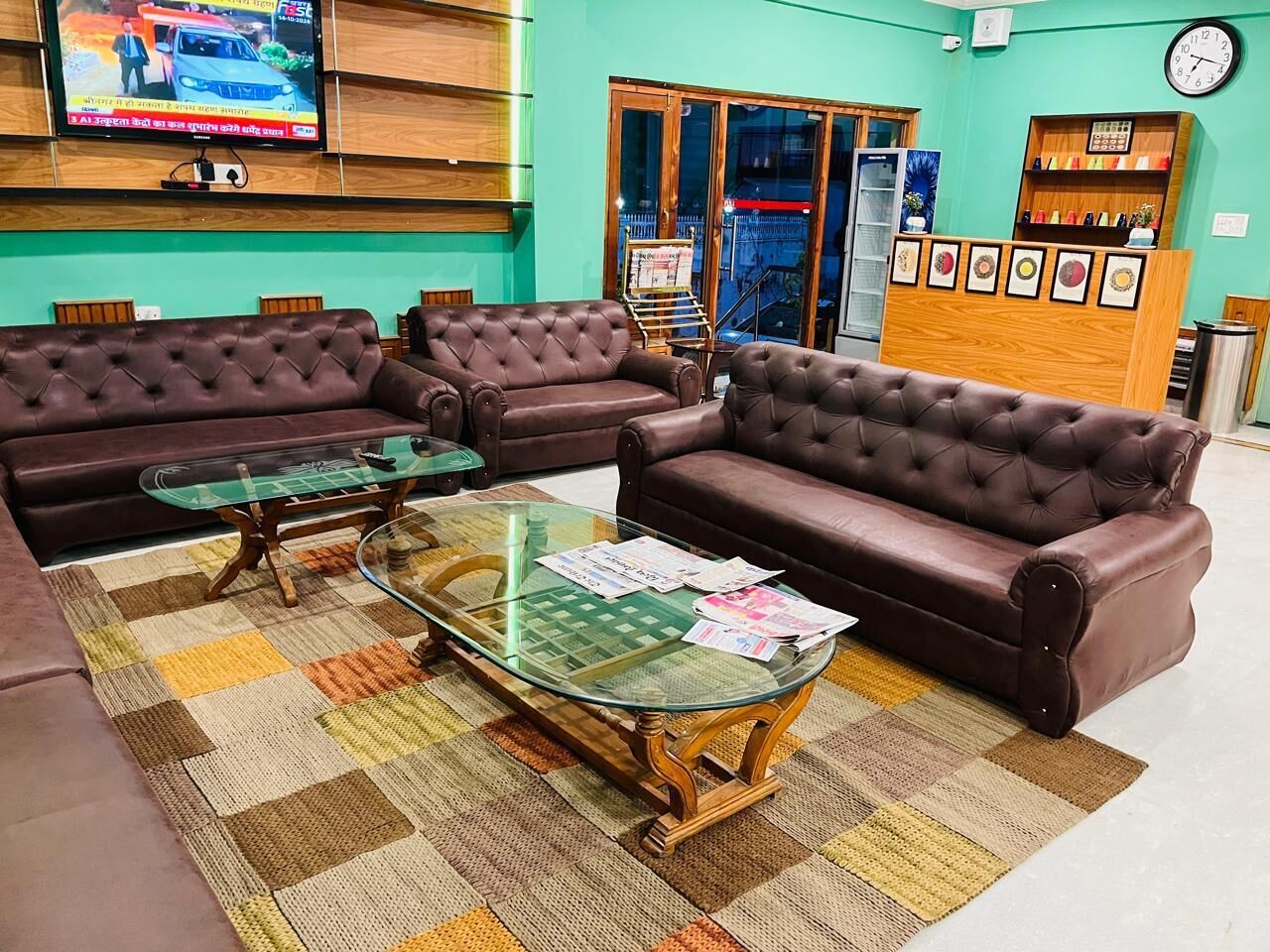 A living room with two brown leather sofas, a glass coffee table, and a checkered rug. A television is mounted on the wall, and there is a wooden reception desk with framed pictures.