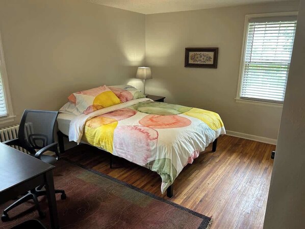 Een slaapkamer met een bed, een bureau, een stoel, een lamp en een raam met jaloezieën.