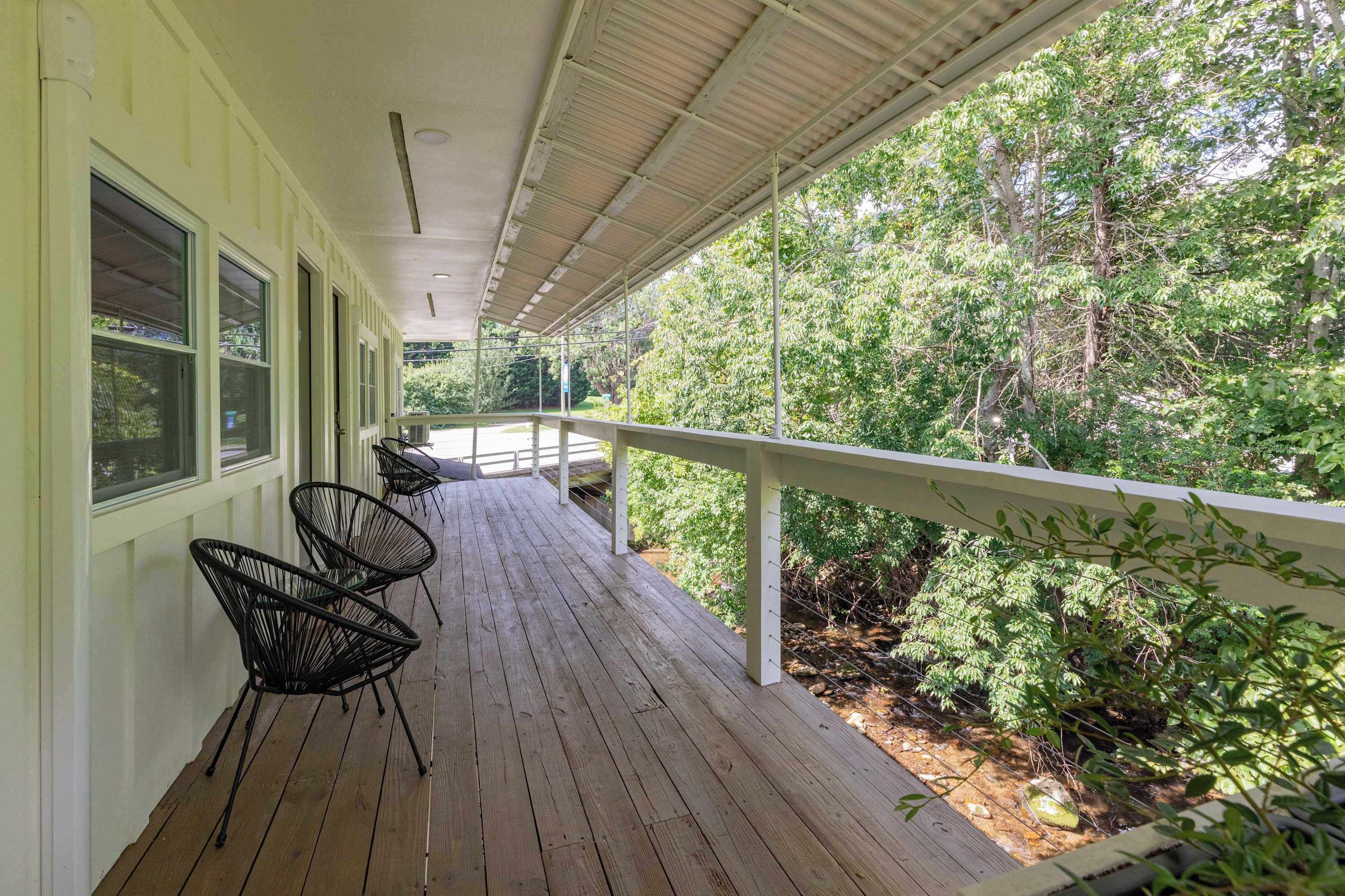 A covered porch with a wooden floor, metal chairs, and a railing overlooking a wooded area.