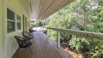 A covered porch with a wooden floor, metal chairs, and a railing overlooking a wooded area.