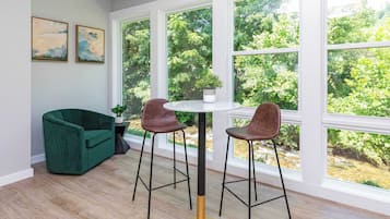 A room with a green sofa, two bar stools, a round table, and large windows with a view of trees.