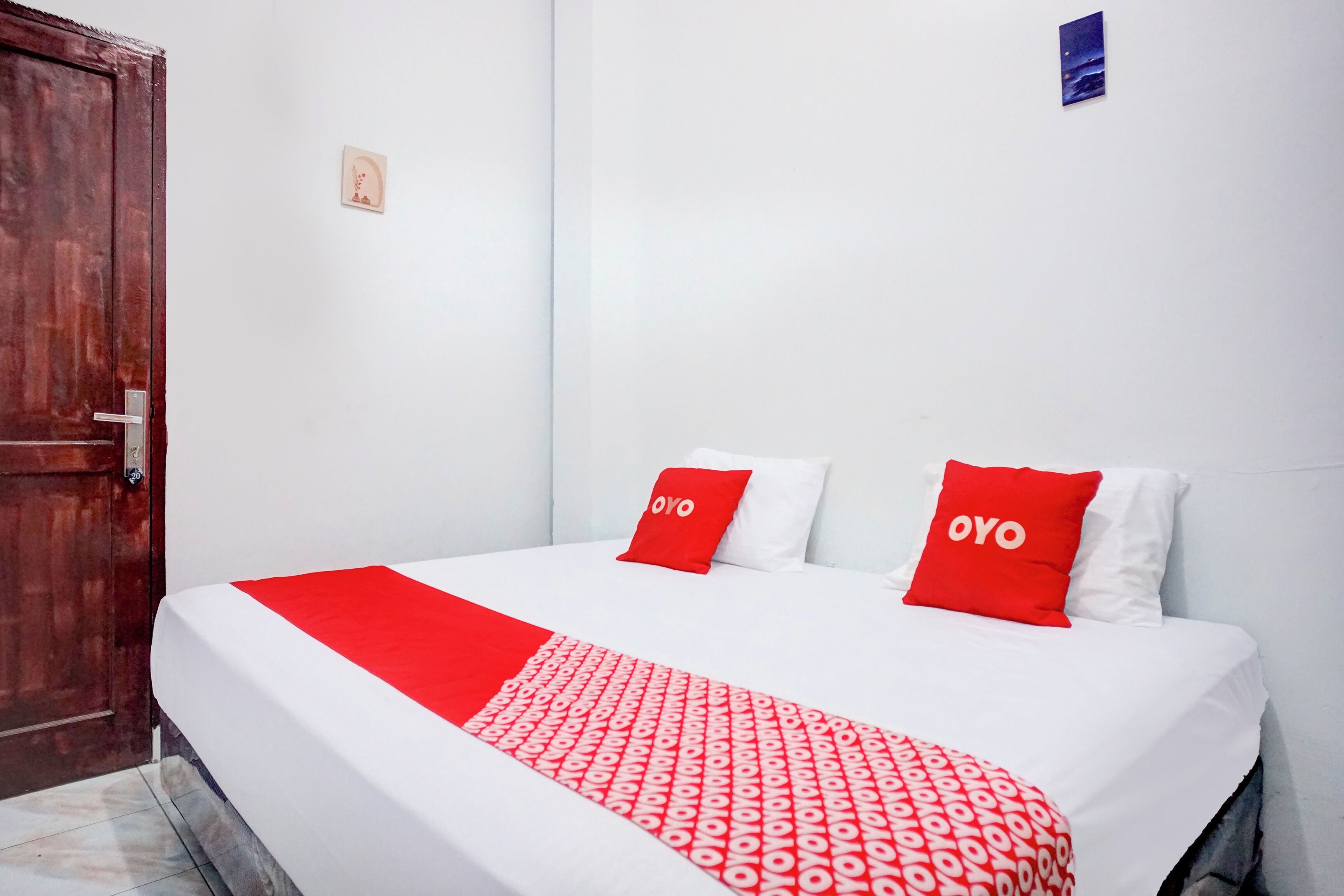 Habitación de hotel con cama, dos almohadas rojas con la palabra "OYO", una colcha blanca y una manta roja con un estampado.