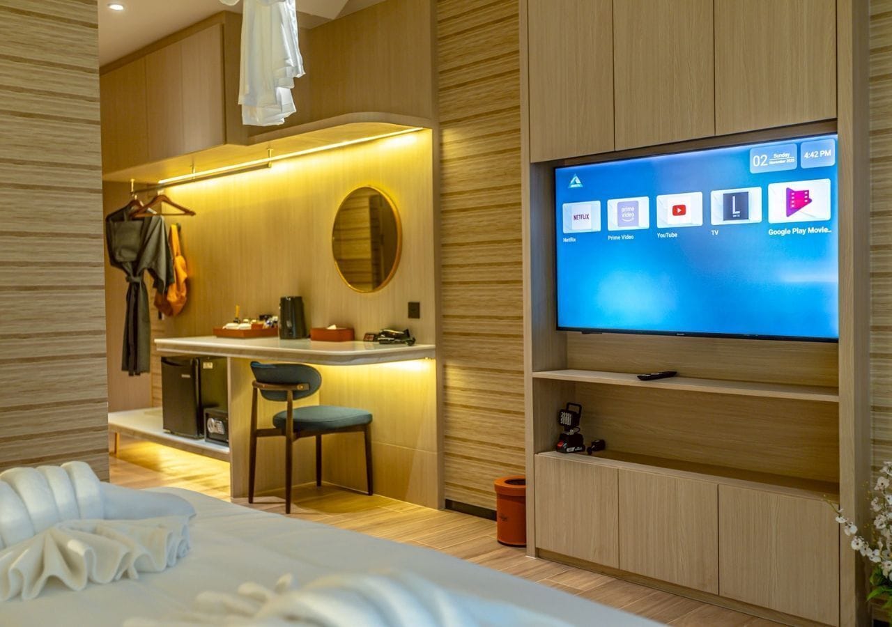 Quarto de hotel moderno com uma cama, uma escrivaninha com cadeira, uma televisão na parede e uma pequena área de cozinha.