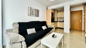 Una sala de estar con un sofá color beige, un plaid negro, una mesa de centro blanca y una cocina al fondo.