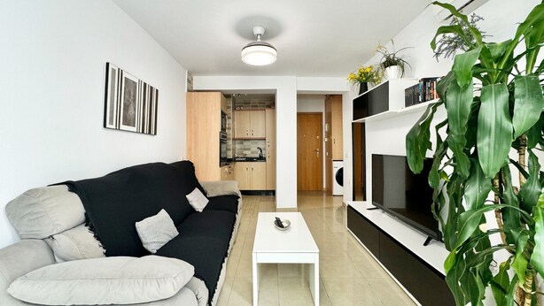 Una sala de estar moderna con un sofá blanco y negro, una mesa de centro blanca y un televisor sobre un soporte negro. Se observa una cocina al fondo con armarios de madera y un microondas.