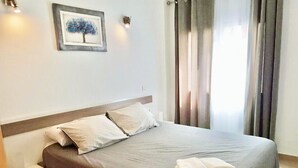 Una cama bien hecha con colcha gris y almohadas blancas, una toalla blanca doblada sobre la cama, una mesita de noche con una lámpara y un cuadro en la pared.