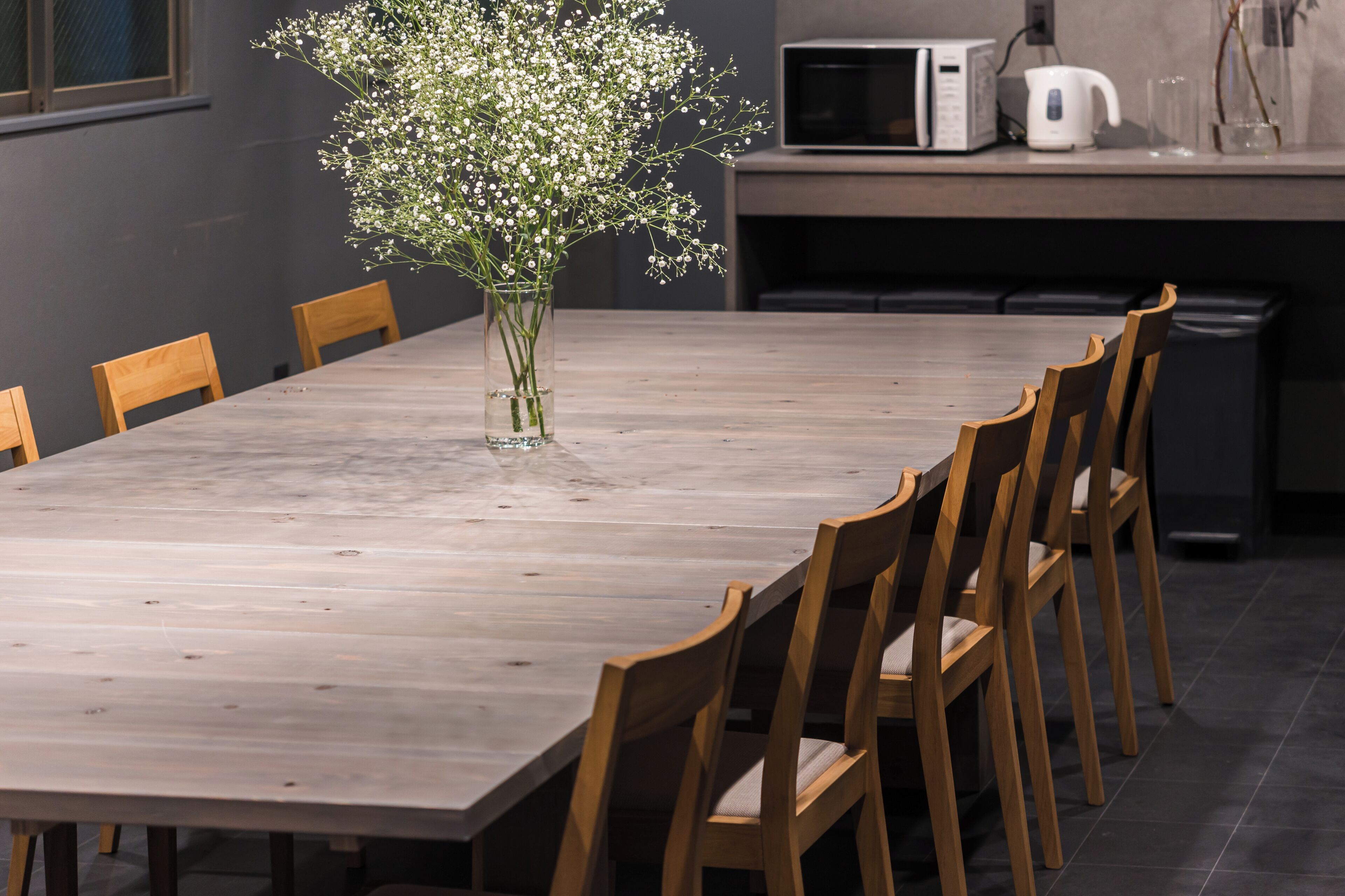 A dining table with chairs, a vase with flowers, a microwave, and a kettle on a kitchen counter.