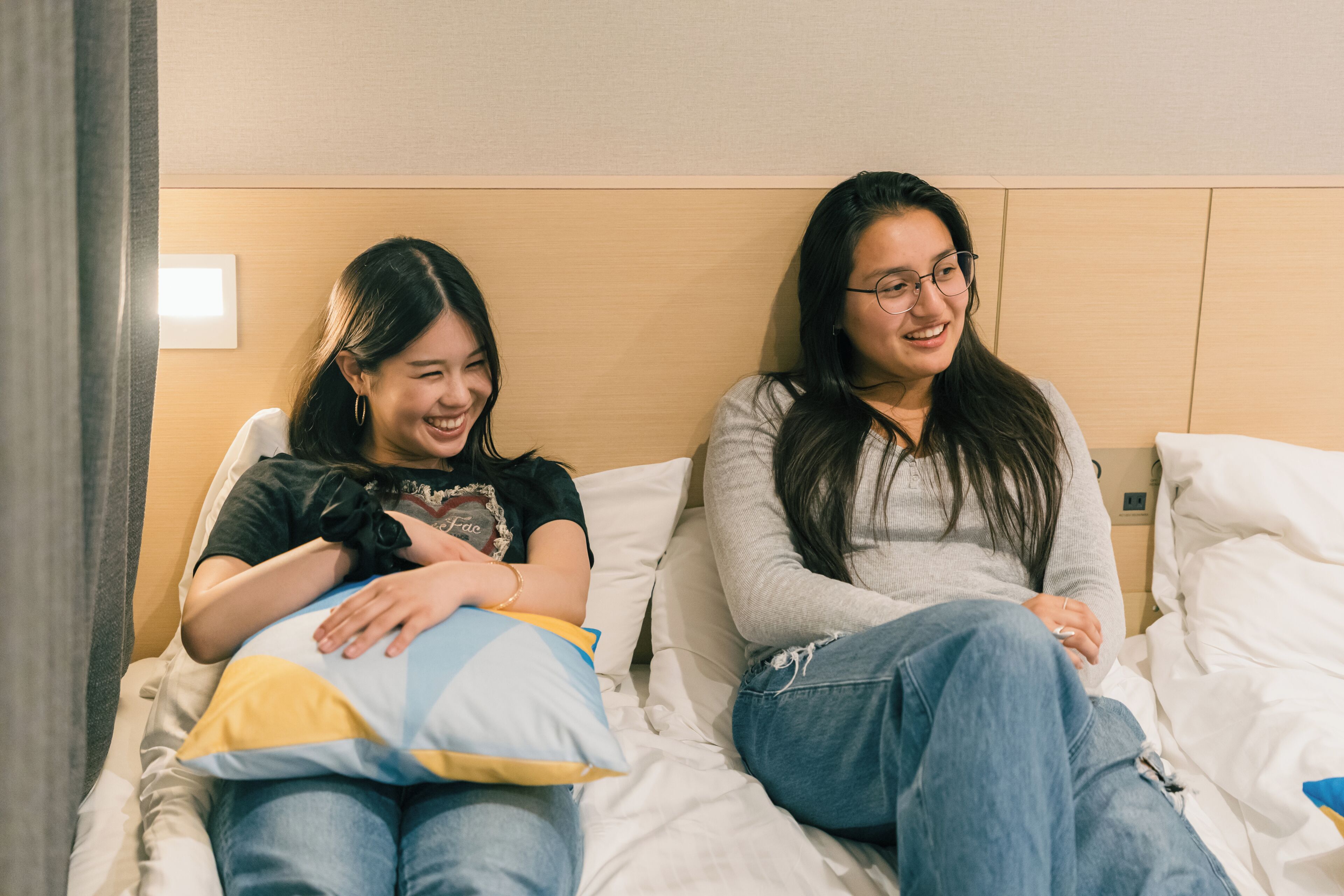 Two individuals sitting on a bed with white bedding, a wooden headboard, and a visible wall socket.